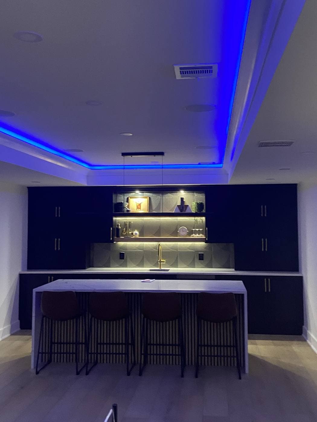 A dark bar with a marble waterfall island, blue cove lighting around a tray ceiling, and warm lit shelving behind the counter.