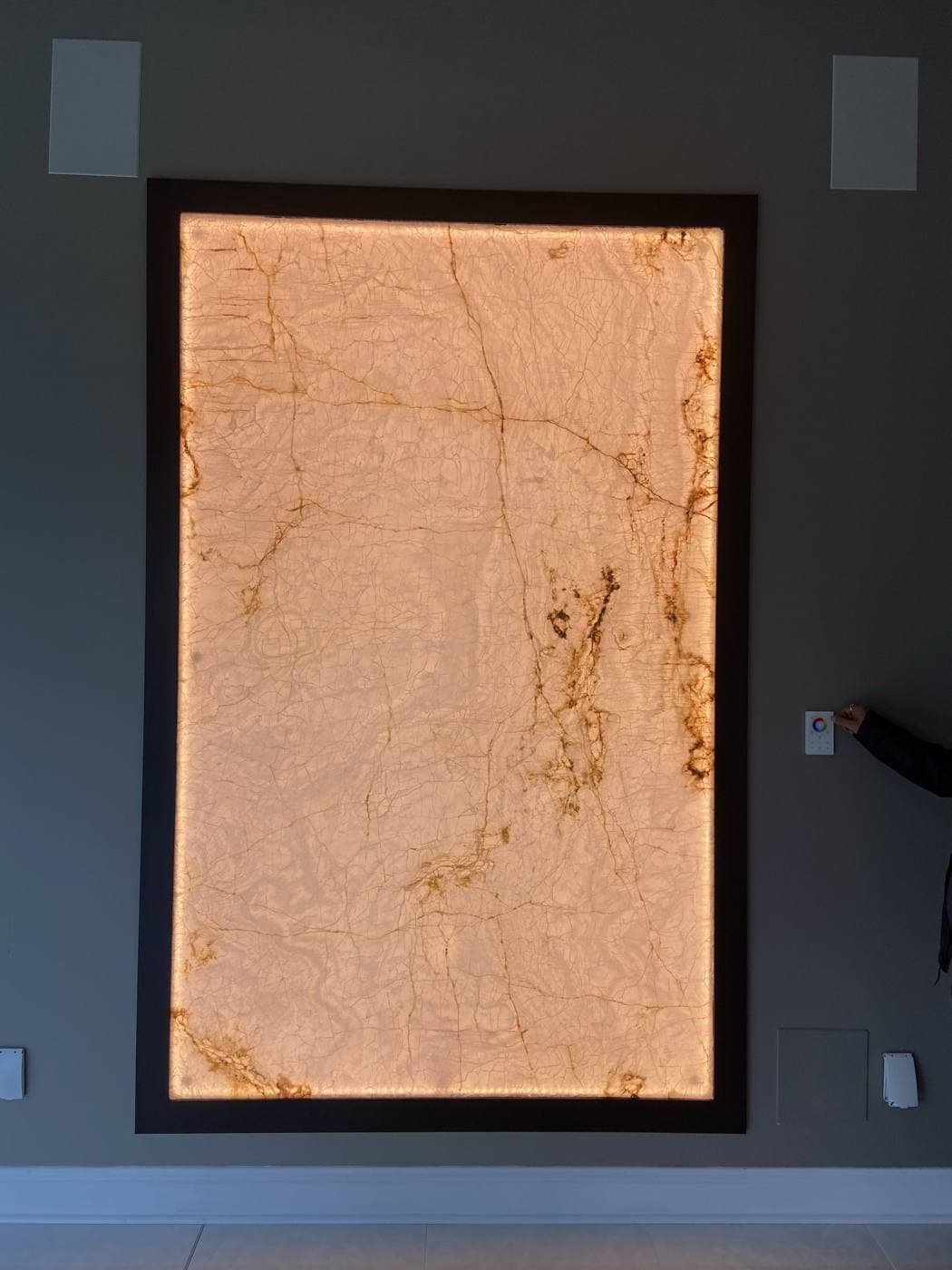 A large backlit stone panel set into a dark wall, glowing warm amber, with a hand adjusting a wall-mounted control beside it.